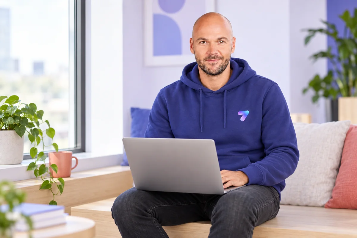 Tobias Hieb sitting with a laptop on his lap, smiling into the camera, in a bright room with plants and a blue hoodie that has the SevenMail 7 on it